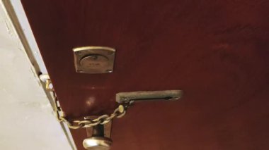 Yan görüntüde kapalı sahne, sabit kamera, yakın çekim, evin kapısında zincir kilidi açmaya çalışan bir eli gösteriyor..