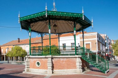 Mavi gökyüzünün altındaki küçük bir kasaba meydanında klasik yeşil demirden bir bando tezgahının ön görüntüsü toplumu ve geleneği simgeler, penaranda de bracamonte, İspanya