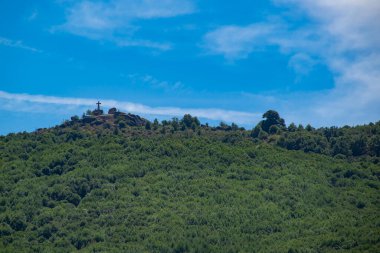 Yoğun ağaçlar ve bitki örtüsüyle kaplı yemyeşil bir tepenin alçak açılı görüntüsü mavi bir gökyüzüne karşı. Uzakta küçük bir haç ya da anıt görülebilir.