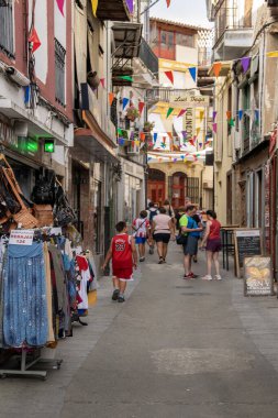 Hervas, İspanya: 19 Ağustos 2023: Turistler renkli bayraklar, hervalar, İspanya 'da dolaşırken, yerel dükkanların sergilendiği dar ve işlek bir caddeye bakarken dikey perspektif görüntüsü