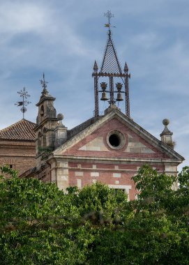A wide shot of an old brick church with a bell gable standing behind a canopy of lush green trees under a cloudy sky, hervas, spain