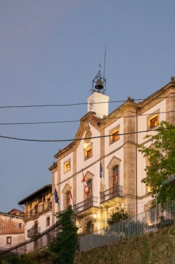 Candelario, İspanya: 19 Ağustos 2023: Alacakaranlıkta park etmiş arabalarla eğimli bir caddenin üzerinde yükselen aydınlık bir kilisenin geniş açılı görüntüsü; Candelario; İspanya;;