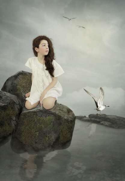 Little girl with long hair in white nightgown sitting on the rock above the water before dawn and seagulls