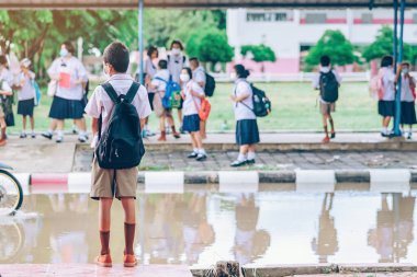 Erkek ilkokul öğrencisi Coronavirus 'u (Covid-19) önlemek için yüz maskesi takıyor ailesinin okuldan sonra eve dönmesini bekliyor ve okul kapısının önünde yağmur duruyor.