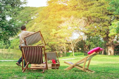 Erkek garsonun arka görüntüsü müşteriler için güverte sandalyeleri hazırlayıp bahçede dinlenmelerini sağlar. Yeşil bir çevrede yaz tatili. Bahçedeki iskemlede rahatlarken mutlu ol. Açık hava eğlencesi..