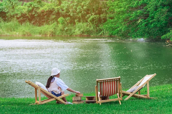 Back view of Asian woman with white hat sitting on garden chair near pond in garden. Summer vacation in green surroundings. Happy person outdoors relaxing on deck chair in garden. Outdoor leisure.