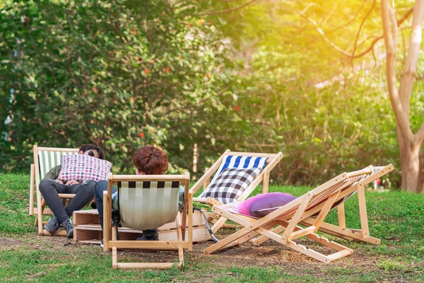 Back view of man and friend sit and napping to relax together on garden chair in garden. Summer vacation in green surroundings. Happy person outdoors relaxing on deck chair in garden. Outdoor leisure.