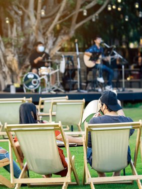 Asyalı genç çiftin arka planı, arka planda müzikal performansın bulanık görüntüsüyle çimenlikte akşam yemeği için masalarla beyaz güverte sandalyelerinde rahatlamak için otururken, gölgeli yeşil çimlerle çevrilidir..