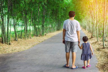 Asyalı babanın el ele tutuşup bambu bahçesinde yürüyen güzel kızı tutuşu. Baba ve kızı parkta birlikte yürüyorlar. Mutlu aile, yeşil doğada birlikte vakit geçiriyor..