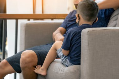 Asyalı bir çocuğun yüz maskesi takıp koltukta akıllı telefon oynaması. Cep telefonu olan küçük çocuk kafede koruyucu maske takıyor. Hastalık salgını sırasında halka açık yerleri ziyaret etme kavramı. Yeni normal.