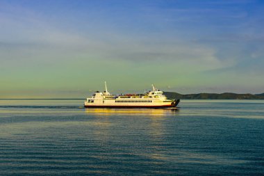 Durgun suda seyreden yolcu feribotu görüntüsü. Beyaz yolcu feribotu kristal berrak turkuaz sularda yol alıyor dramatik adaları ve mavi gökyüzünün altında görünen dağ manzarasıyla..