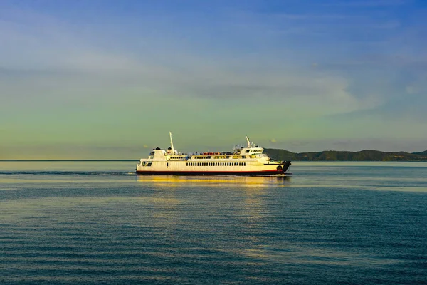 Durgun suda seyreden yolcu feribotu görüntüsü. Beyaz yolcu feribotu kristal berrak turkuaz sularda yol alıyor dramatik adaları ve mavi gökyüzünün altında görünen dağ manzarasıyla..