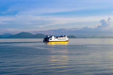 Durgun suda seyreden yolcu feribotu görüntüsü. Beyaz yolcu feribotu kristal berrak turkuaz sularda yol alıyor dramatik adaları ve mavi gökyüzünün altında görünen dağ manzarasıyla..