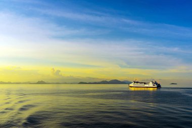 Durgun suda seyreden yolcu feribotu görüntüsü. Beyaz yolcu feribotu kristal berrak turkuaz sularda yol alıyor dramatik adaları ve mavi gökyüzünün altında görünen dağ manzarasıyla..