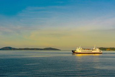 Durgun suda seyreden yolcu feribotu görüntüsü. Beyaz yolcu feribotu kristal berrak turkuaz sularda yol alıyor dramatik adaları ve mavi gökyüzünün altında görünen dağ manzarasıyla..