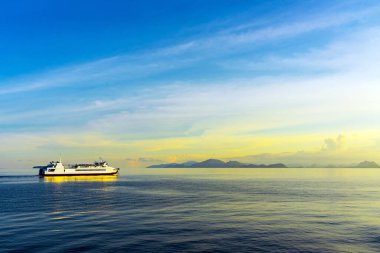 Durgun suda seyreden yolcu feribotu görüntüsü. Beyaz yolcu feribotu kristal berrak turkuaz sularda yol alıyor dramatik adaları ve mavi gökyüzünün altında görünen dağ manzarasıyla..
