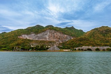 Patlayan Dağlar. Bulutlu gökyüzünün altında nehir tepe taş ocağı manzarası. Dağları ve bulutlu arka planı olan nehir kıyısındaki taş ocağının manzarası. Dağ manzarasına çevresel etki.