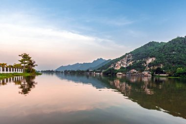 Patlayan Dağlar. Bulutlu gökyüzünün altında nehir tepe taş ocağı manzarası. Dağları ve bulutlu arka planı olan nehir kıyısındaki taş ocağının manzarası. Dağ manzarasına çevresel etki.