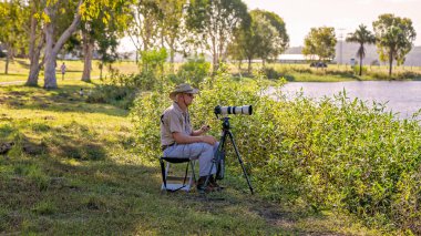 Mackay, Queensland, Australia - January 2021: Male retiree photographer taking photos of birds on a pond in late afternoon sunlight. clipart