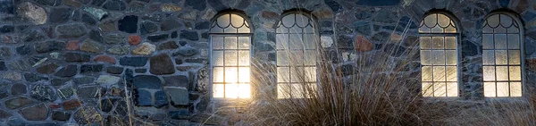 Four windows set into a stone wall with dried grass in front and a light glow shining through