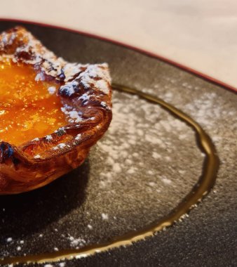 Sıcak Portekiz kremalı tart üzerine krema serpilmiş ve koyu bir tabakta servis edilmiş.