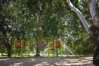 Açık hava parkındaki gölgeli ağaçların arasında bir çocuk parkı.