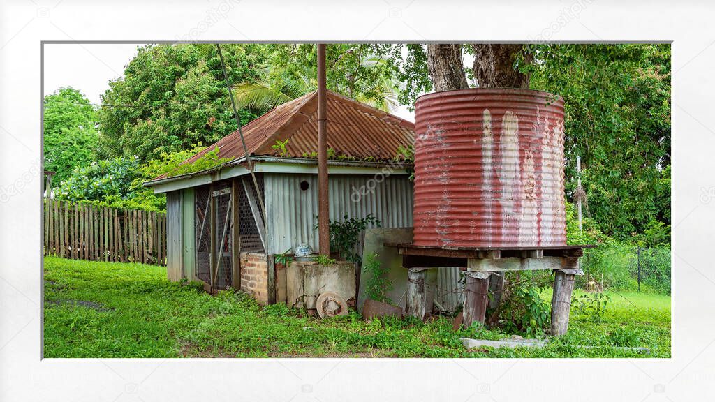 Old shed and water tank in an outdoor backyard after rainfall when the grass and foliage are lush and green, with white mat frame