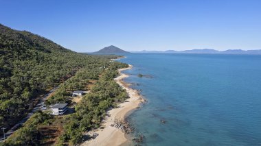 Okyanus üzerinde insansız hava aracı antenleri ve deniz kenarındaki evleri gösteren kayalarla dolu uzun kumlu bir plaj.