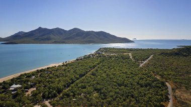 Uzun bir kumlu sahil ve dağ arkaplanı olan bir sahil yerleşim bölgesinin hava manzarası.