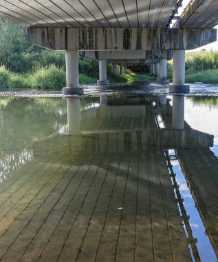 Sığ bir kırsal derenin üzerine inşa edilmiş beton köprünün altında su yansımaları var.