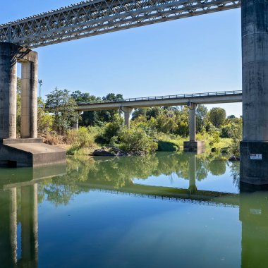 Kırsal bir dereye yayılan demiryolu ve otoyol köprüleri, gölgeler ve su yansımaları sabah ışığında