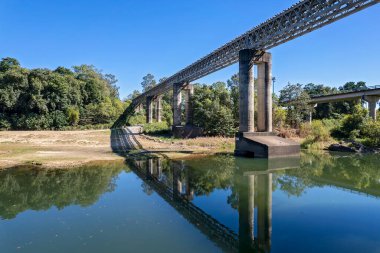 Kırsal bir dereye yayılan demiryolu köprüsü, karayolu köprüsü, gölgeler ve su yansımaları.