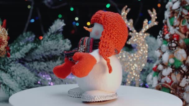 Egy vörös kalapos hóember forog a peronon, a karácsonyi fények hátterében.