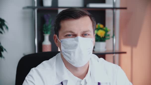 Portrét doktora v masce sedícího v kanceláři. Portrét mladého bělocha s lékařskou obličejovou maskou dívající se do kamery. Lékařský pracovník v ochranné masce. — Stock video