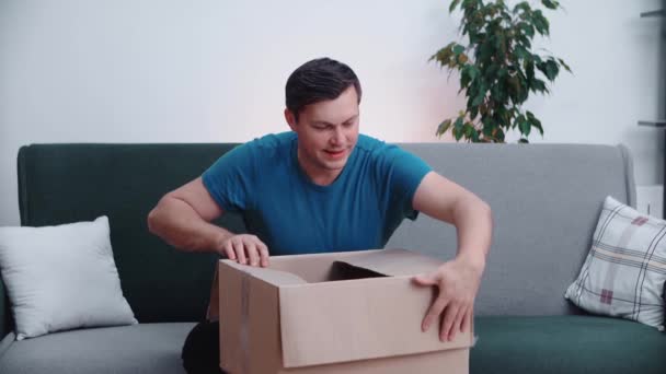 젊은 남자 가집 소파에 앉아 있는 동안 판지 상자를 열었다. 행복 한 소비자는 우편 배달을 하고 구매하는 것으로 만족 한다 — 비디오