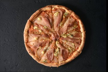 Koyu gri arka planda jambon ve parmesan peynirli yuvarlak pizzanın üst görüntüsü..