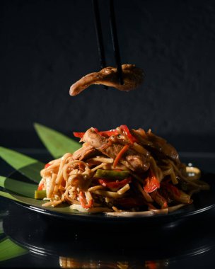 Udon şehriyelerinin seçici odağı domuz parçaları ve siyah tabakta siyah arka planda sebzeler..