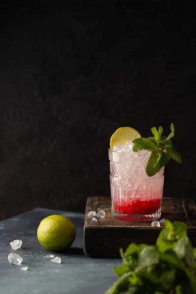 Refreshing cocktail with vodka,soda,berry puree,lime,mint and crushed ice on wooden box and dark background.