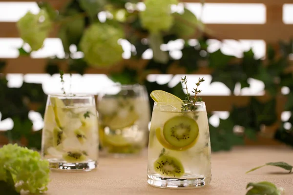 Cocktail rinfrescante o tè freddo con fette fresche di kiwi, rametti di timo, lime e ghiaccio in un fronte di fiori estivi.. — Foto Stock