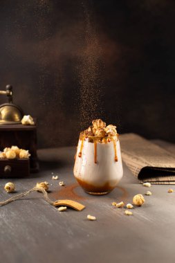 Sweet Milkshake with caramel syrup,cream liqueur,caramel popcorn and chocolate powder on brown background with vintage,manual coffee grinder and falling cocoa powder. clipart