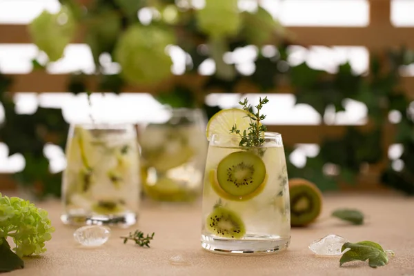 Cocktail rinfrescante o tè freddo con fette fresche di kiwi, rametti di timo, lime e ghiaccio in un fronte di fiori estivi.. — Foto Stock