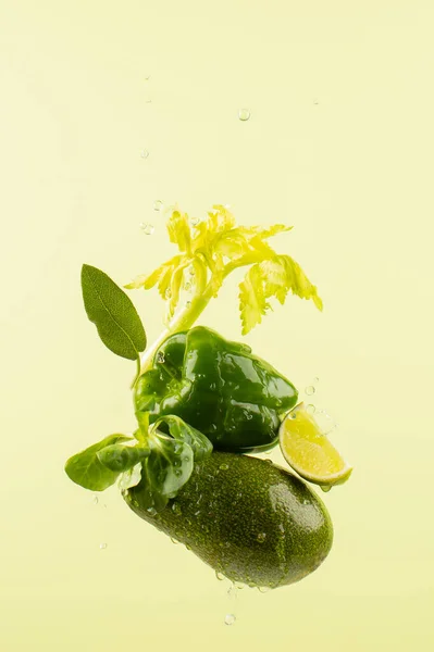 Fresh raw vegetables arranged in a stack:avocado,bell pepper,lime,celery,corn salad and sage leaf on light green background.Flying vegetables with water drops.Healthy and vegan food concept.