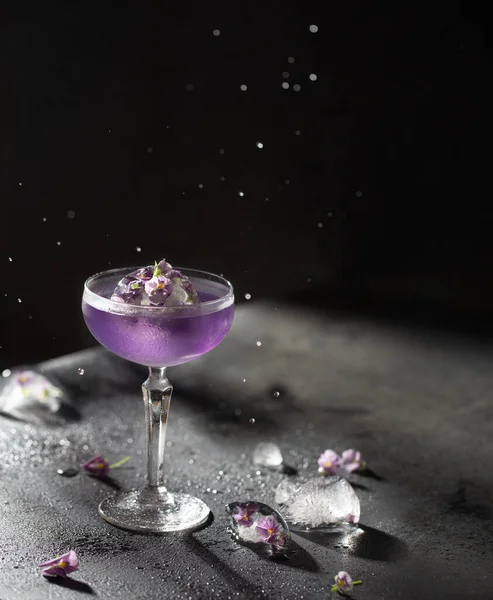 Bellissimo cocktail viola in un bicchiere di cristallo con gin, soda e palla di ghiaccio congelati con fiori delicati su sfondo nero con gocce volanti e ombre dure.. — Foto Stock