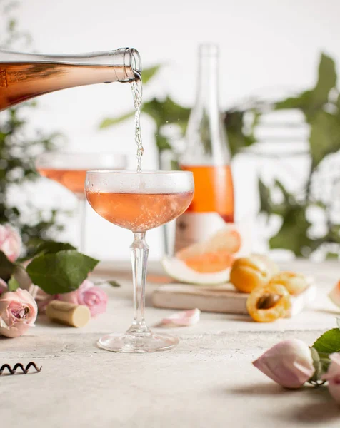 Розовое вино в хрустальных бокалах и бутылка со свежими абрикосами, грейпфрутами, нежными розами и зелеными растениями на белом фоне.. — стоковое фото