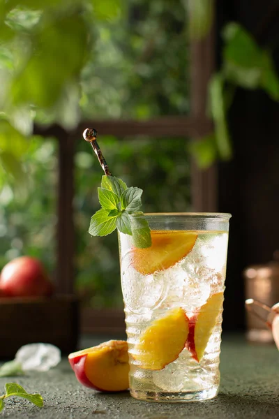 Cocktail rinfrescante o tè freddo con pesche fresche e foglie di menta davanti a una finestra di legno con piante estive verdi.Detox, bevanda sana. — Foto Stock