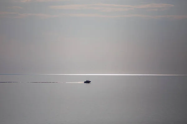 Open sea. A boat is sailing in the distance. The sky almost merges with water. The boat trail is parallel to the horizon. There is copy space.