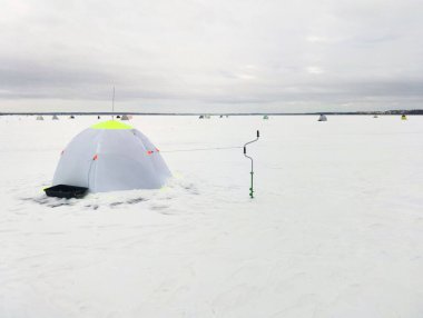 Balıkçıların çadırı ve elle tutulan buz matkabı karla kaplı bir göle karşı balıkçı kulübesinde.