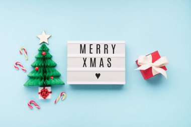 Mutlu Xmas yazıları beyaz ışıklı kutuda, üzerinde yeşil ağaç, mantar ve mavi arka planda kırmızı hediye kutusu var. Düz konum, üst görünüm, kopyalama alanı. Bayram kartı konsepti.