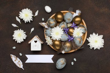 Paskalya bayramı tebrik kartı. Gri mavi altın yumurtalı Paskalya Süslemesi, bıldırcın tüyleri, koyu ahşap arka planda beyaz çiçekler. Paskalya kartı konseptin kutlu olsun. Boşluğu kopyala, üst görünüm.