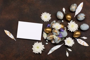 Paskalya bayramı tebrik kartı. Gri mavi altın yumurtalı Paskalya Süslemesi, bıldırcın tüyleri, koyu ahşap arka planda beyaz çiçekler. Paskalya kartı konseptin kutlu olsun. Boşluğu kopyala, üst görünüm.